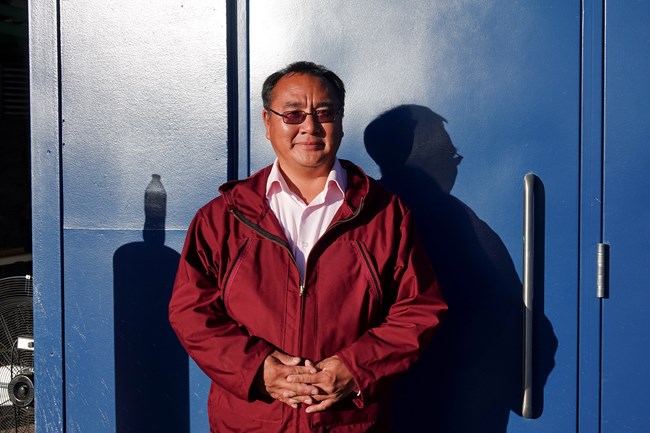 A smiling Alaska Native in a red jacket against the backdrop of a blue building.