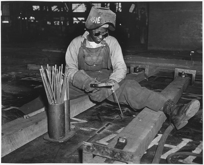 African American woman wearing overalls, work boots, thick gloves, dark goggles, and a raised welding helmet looks down at a piece of equipment in her hand. She is sitting with legs splayed out as she works on wooden beams below her.