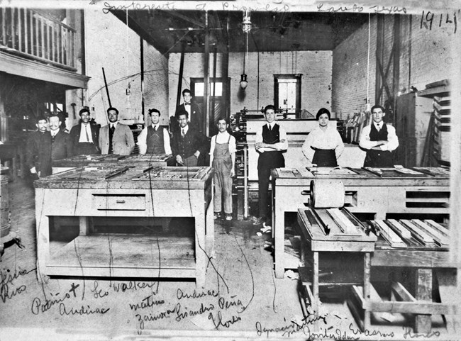 Print shop of El Progreso newspaper Group of 11 people standing in a room with exposed brick walls and concrete floors surrounded by newspaper printing machinery