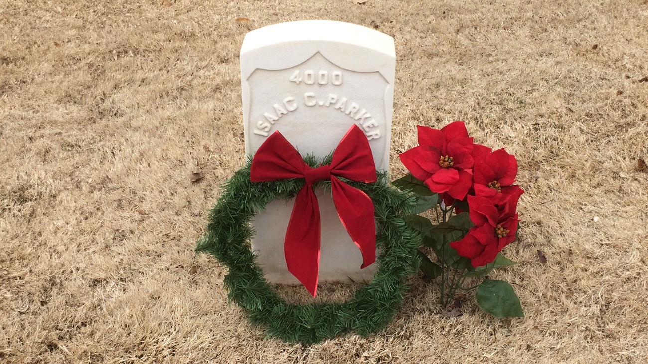 Judge Parker's headstone at the Fort Smith National Cemetery A white headstone in brown grass with a wreath and poinsettia plant laid beside it. Text reads, "4000, Isaac C. Parker."