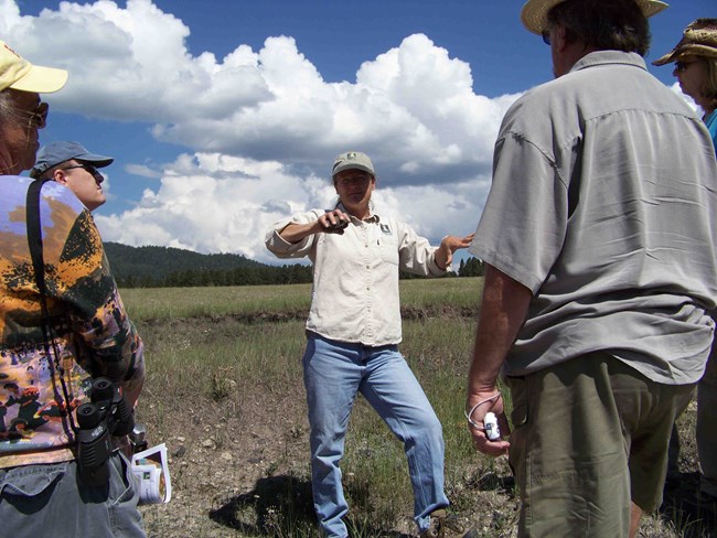 A woman addresses a group of people in a grassland.