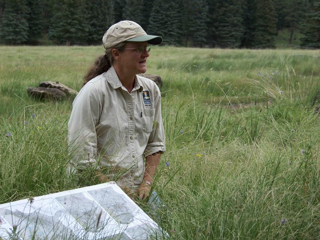 A scientist kneels in the grass while addressing a group of people in a large valley.