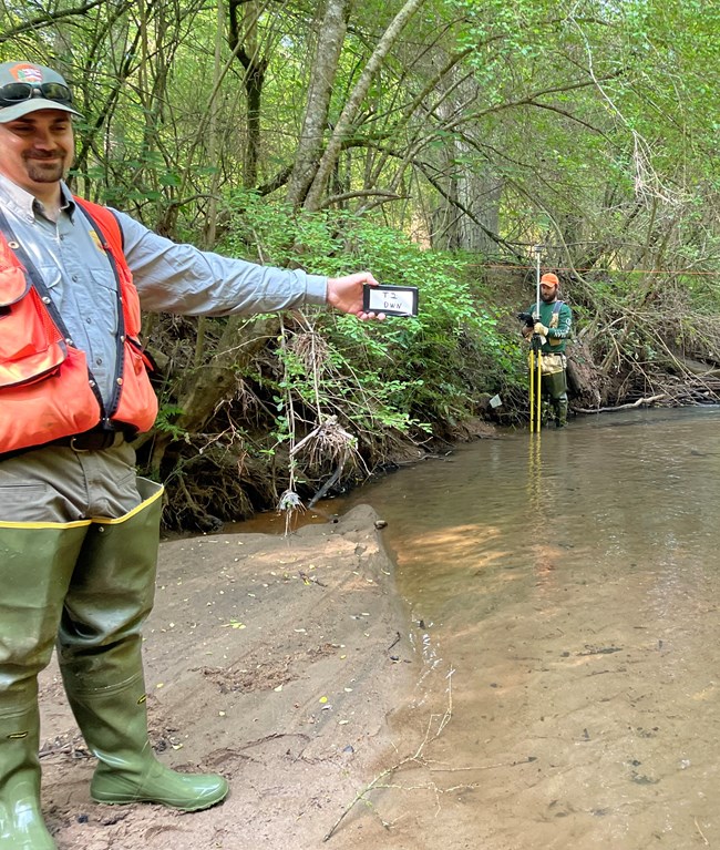 Man wearing orange vest holding a sign next to a stream. Man with survey equipment in background