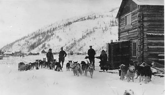 Black and white photograph looking north at the Historic Kantishna Roadhouse with the Kantishna Hills in the background, taken around 1921. In front of the roadhouse are three dog teams and four unidentifiable people.