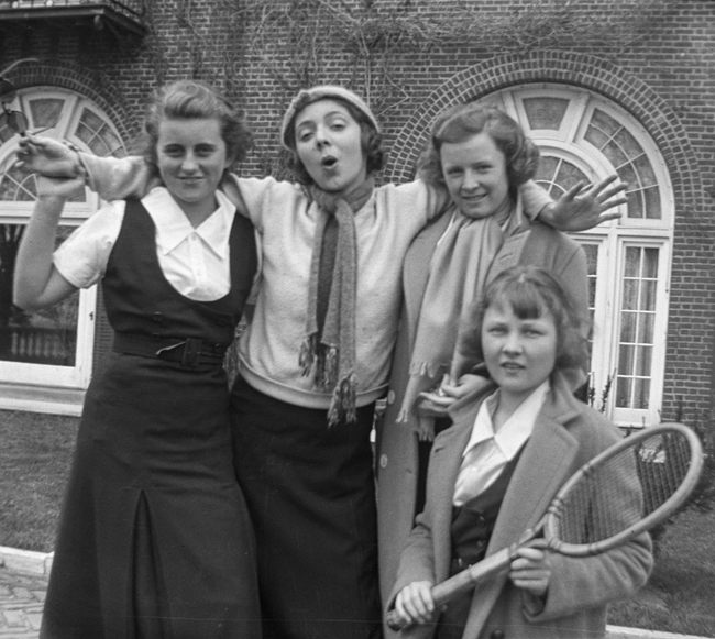 Kick with Noroton friends A black and white photo of four adolescent girls in skirts and curled hair posing in front of a brick building. One stands in center, her arms around the others’ shoulders, hand held by the girl left of her, and another is in front with a tennis racket.