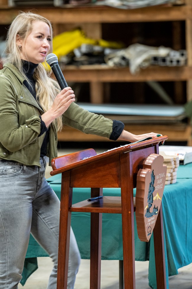 Woman in jeans and a green jacket stands at a lectern and speaks into a stick microphone