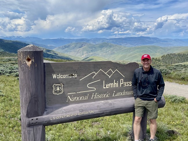 A man stands next to a sign reading "Lemhi Pass National Historic Landmark." The background is lush green hills and a blue sky with fluffy clouds