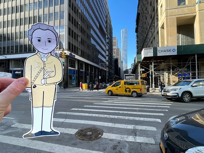 A cartoon illustration cutout of Alexander Hamilton is held against a backdrop of a busy city street.