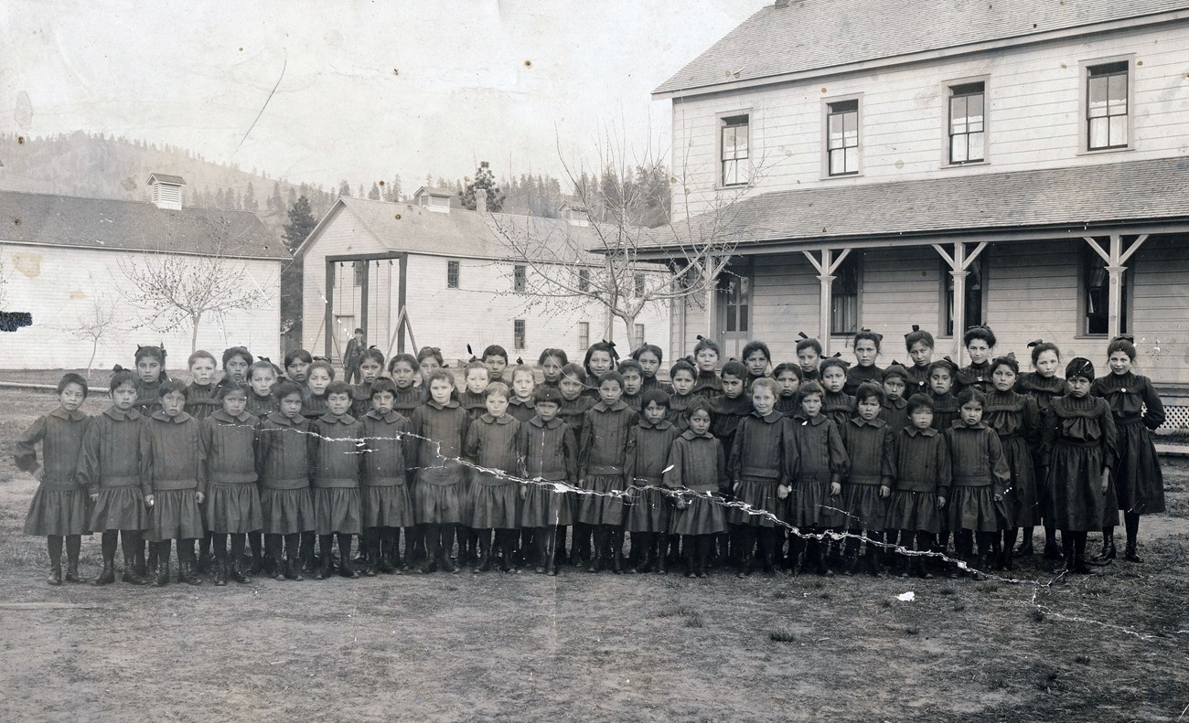 Black and white photograph of several rows of identically dressed Indigenous students standing in front of buildings