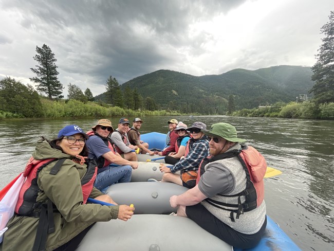 Eight people wearing hats sit on a boat in a river. They are surrounded by lush green hills, trees, and a cloudy sky