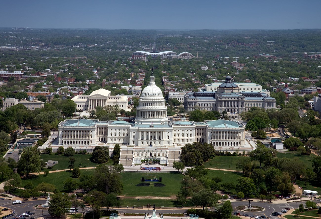 An aerial view of the U.S. Capitol building, it's white dome and flat lower roofs surrounded by trees in downtown Washington D.C.