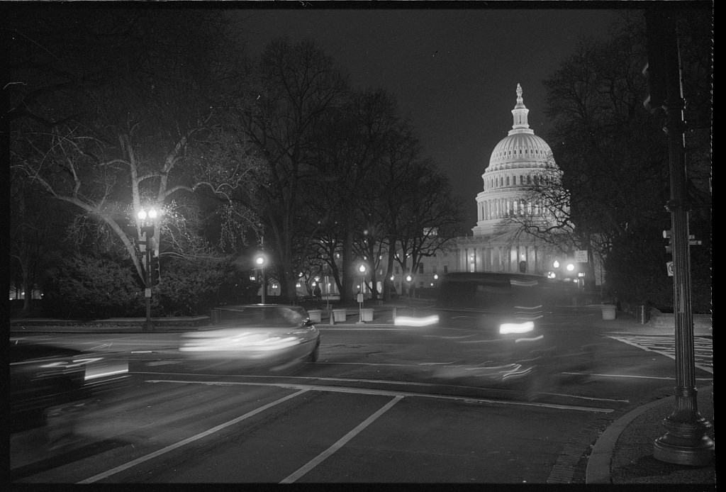 A street view looking toward the U.S. Capitol building at night, the headlights of a passing car bright on the city street