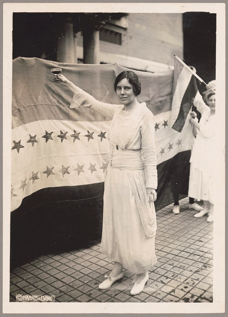 Alice Paul raises a glass in front of a women's suffrage ratification flag with sewn stars