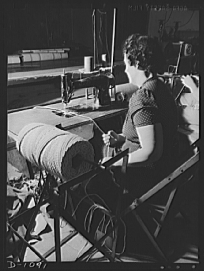 Black and white photo of a white woman with her back to the camera at a sewing machine. A spool of thread in the foreground.