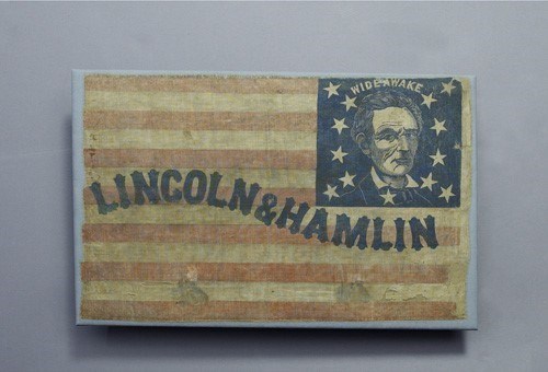 A faded print of a campaign flag. “Lincoln & Hamlin” is printed in blue text over red and white stripes. Lincoln’s face is rendered in a blue square with stars in the upper right corner. Above his head is printed the word “wideawake.”