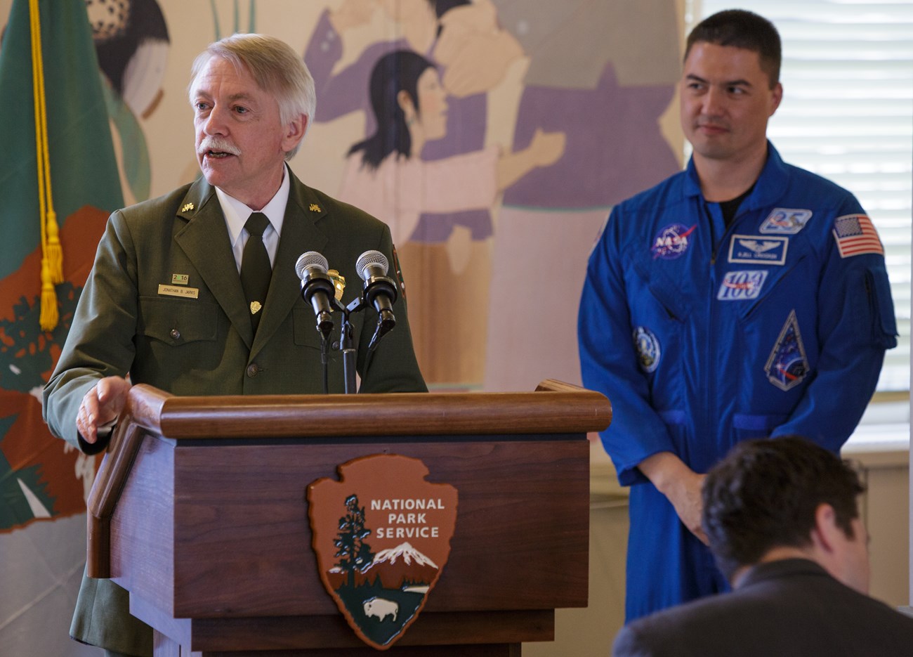 Director Jarvis in his NPS uniforms stands at a podium as Astronaut Kjell Lindgren, in a blue flight suit, looks on.