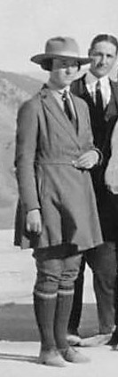 A woman wearing a wide brimmed hat, a long belted coat and knee pants with knee socks.