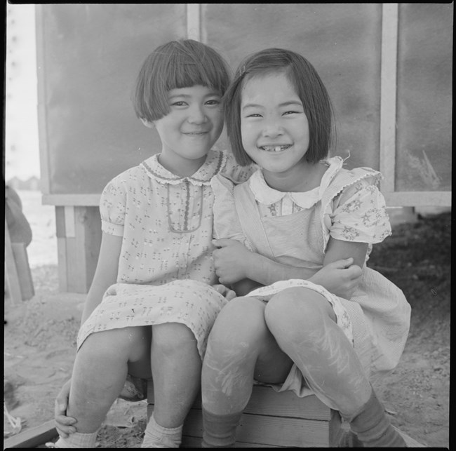Black and white photo two young girls of Japanese ancestry smiling at the camera sitting on a box
