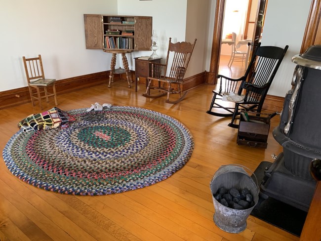 A living room with a large colorful carpet in the middle surrounded by a wood stove, book shelf, and three wooden chairs.
