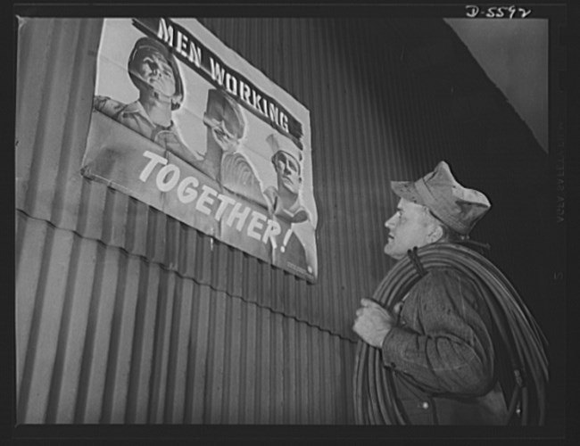 Black and white photo of a white man in an engineer cap carrying hose looking at a wartime poster