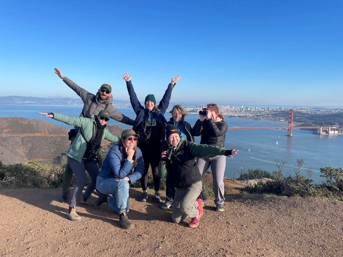 Raptor observatory volunteers. 7 Golden Gate Raptor Observatory volunteers with binoculars posing in front of the Golden Gate Bridge on a hill on a clear day with the cityscape behind them.