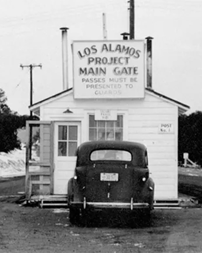 Black and white photo of a small wooden checking station with a 1940s car at front. A sign on the station reads "Los Alamos Project Main Gate. Passes must be presented to guards."