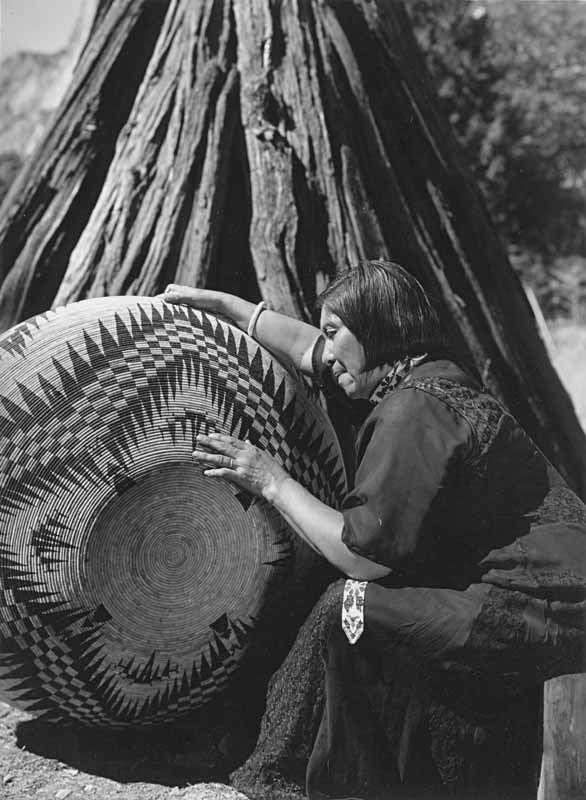 Indigenous woman leans over massive woven basket, sitting in from of umacha