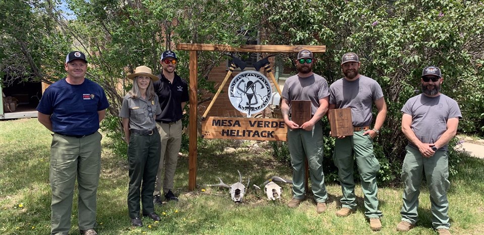 Five men and one woman stand surrounding the Mesa Verde Helitack sign; two men hold awards.