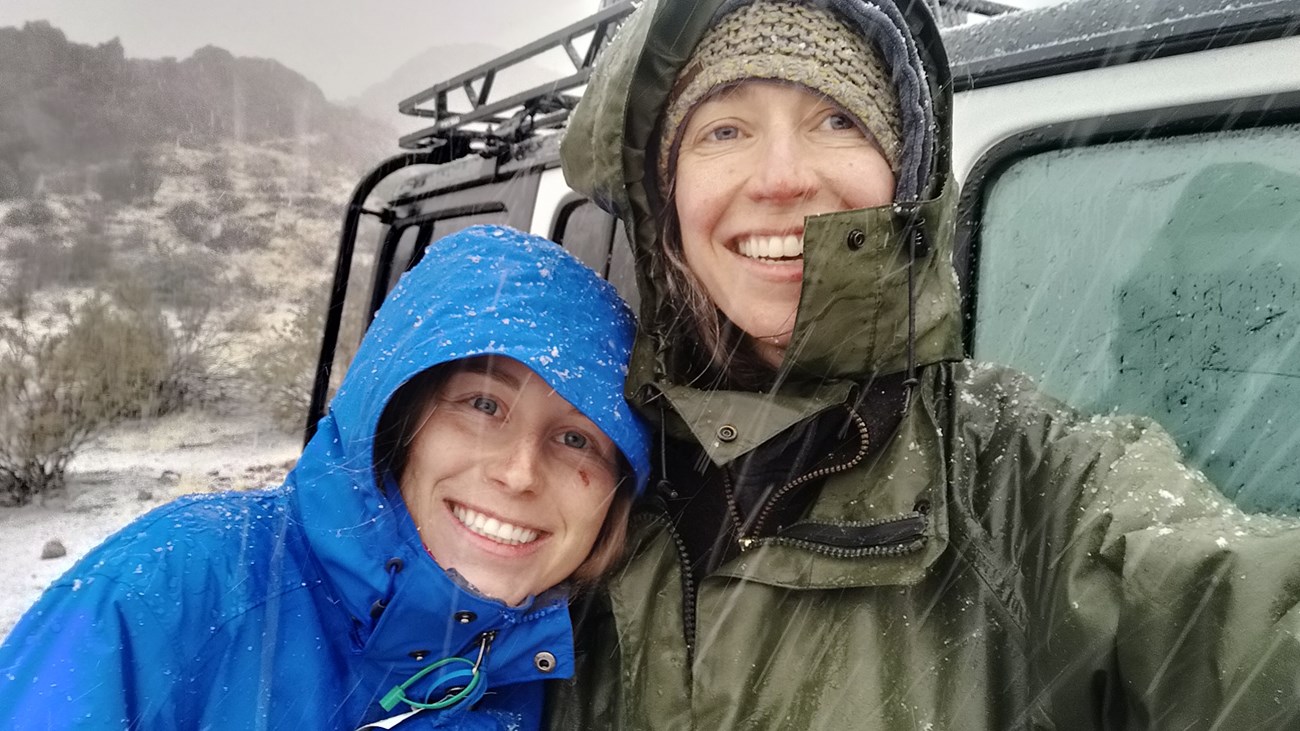 Selfie of two women wearing hooded raincoats smiling at camera in snowfall.