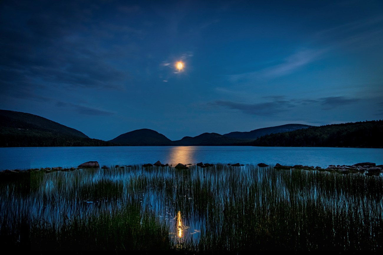 Night photo with dark blue sky, silhouette of mountains in distance, and moon hanging in center with reflections in water below