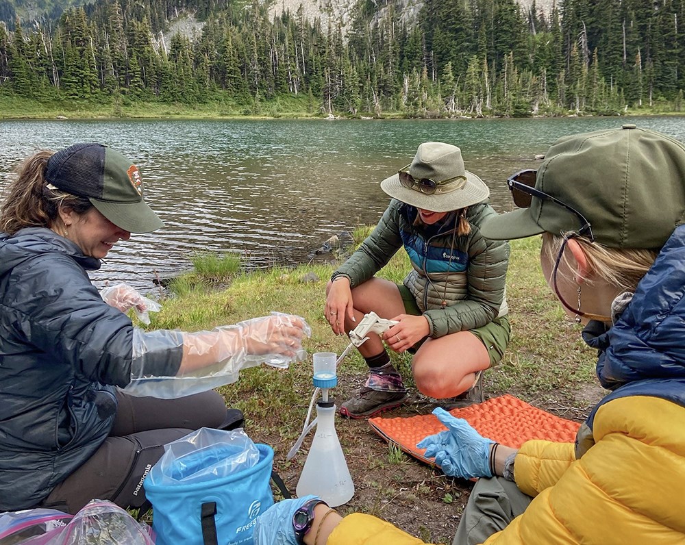 Three people in field clothes sit at the edge of a scenic mountain lake with a plastic flask, a hand pump, and other scientific equipment.