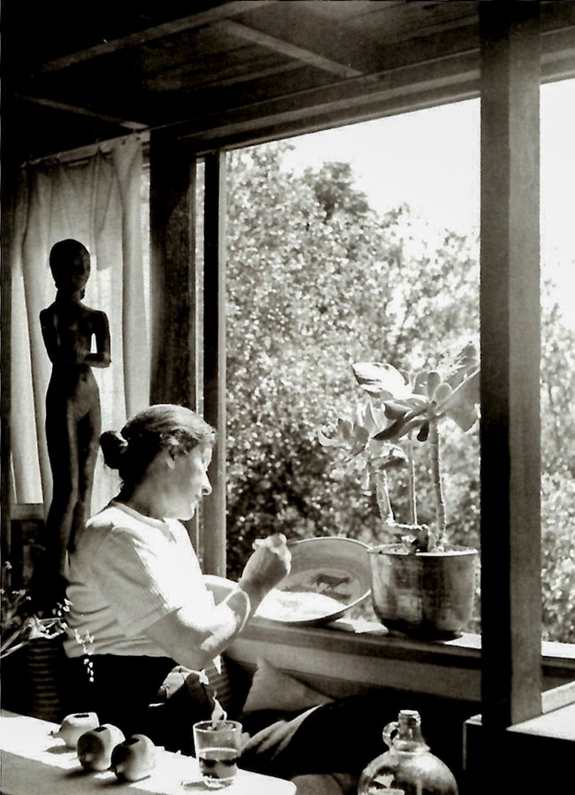 Woman sits at window, paint brush in one hand and clay bowl in another.