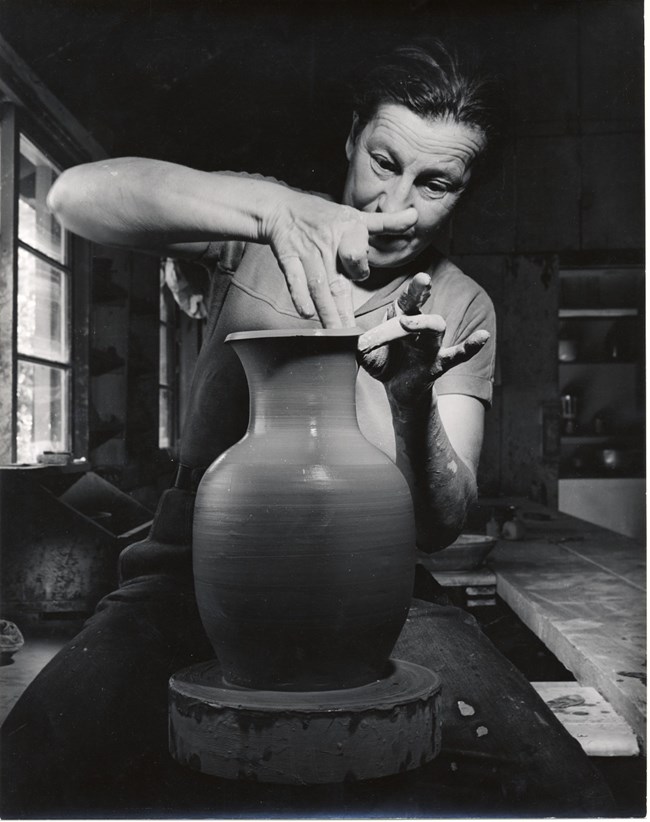 Woman concentrates, using tips of fingers to shape the rim of clay vase on potter wheel.
