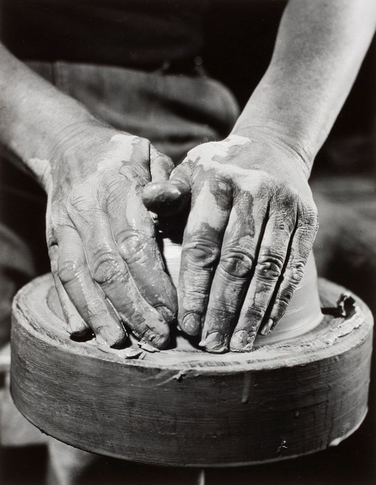 Two muddy hands form ball of clay on potter's wheel.