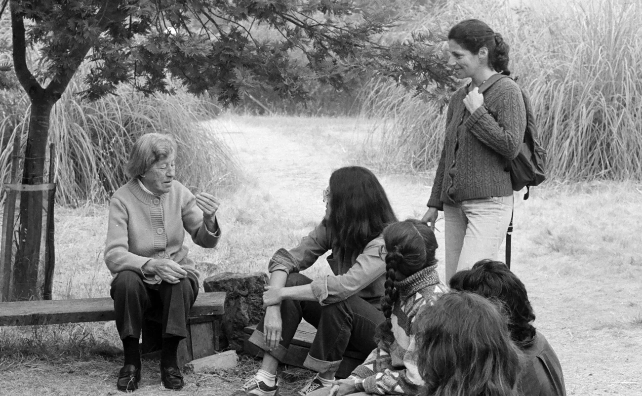 Marguerite with students, ca.1970s Older woman in sweater sits under tree, talking to young students who surround her