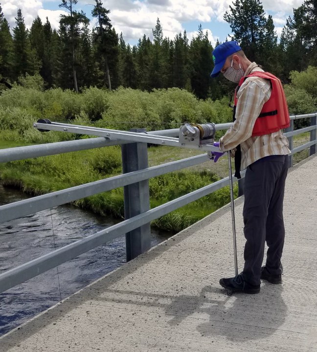 A person on a bridge operating a metal bridge board that has a reel with a sampler hooked to it