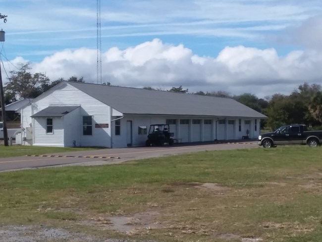 Maintenance HQ A long white building with seven garage bays on a cloudy day is pictured with a golf cart and pickup truck parked by a road to the right of the building. Grass can be seen in the foregorund.