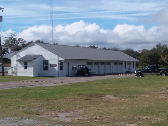 A long white building with seven garage bays on a cloudy day is pictured with a golf cart and pickup truck parked by a road to the right of the building. Grass can be seen in the foregorund.