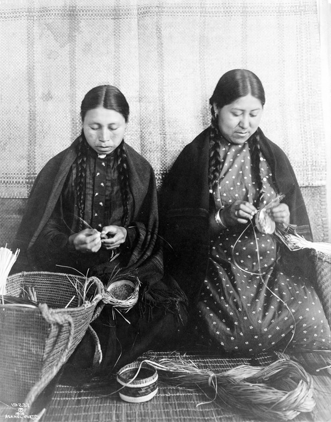 Two women in shawls and long braided pigtails kneel, weaving fibers into baskets.