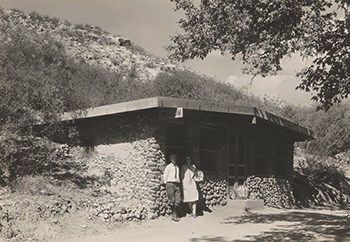 A man and woman stand together in the shade of a stone building at the base of a dusty mountain.