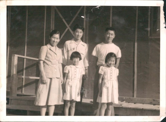 Black and white photo of a Japanese American family: a mother, her two adult sons, and young twin girls. They pose in front of a dark wooden building.