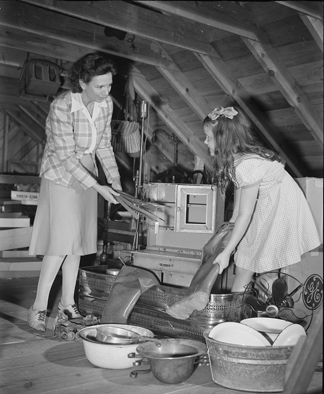 “School children get in the scrap.” Photo by Howard Liberman, Office of War Information, October 1942. Collection of the Library of Congress (https://lccn.loc.gov/2017693935). Black and white photo of two white girls in an attic. They are collecting metal items like buckets, roller skates, and fans as well as rubber boots.
