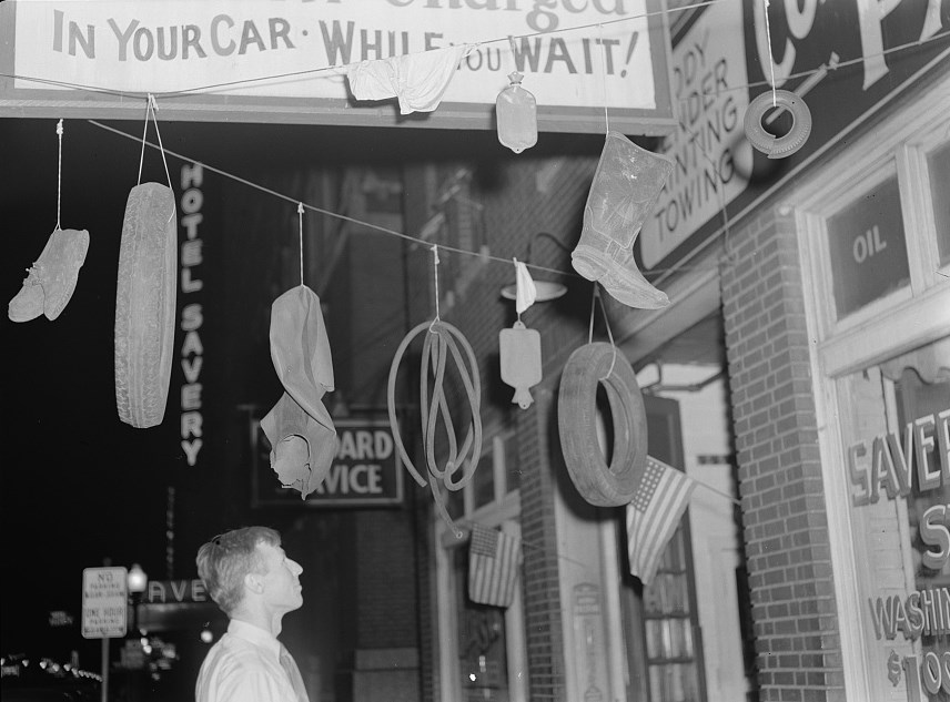 “Salvage.” Photo by Ann Rosener, Office of War Information, September 1942. Collection of the Library of Congress (https://lccn.loc.gov/2017693664). Black and white photo of the exterior of a service center. A white man looks up at rubber items hanging from ropes that have been strung up. They include a tire, rubber boots, a hot water bottle, and a hose.