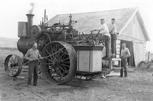 Black and white photo of a large tractor two men stand next to it and two kids are standing on the back