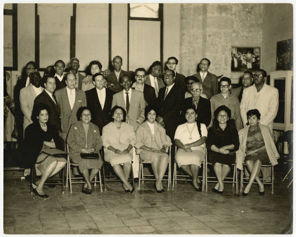 Members of the Orquesta Anacaona pose for a group photograph while sitting in chairs. Others, including men, stand behind them.