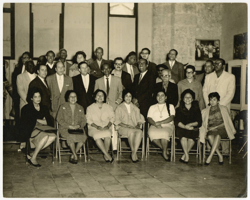 Members of the Orquesta Anacaona Members of the Orquesta Anacaona pose for a group photograph while sitting in chairs. Others, including men, stand behind them.