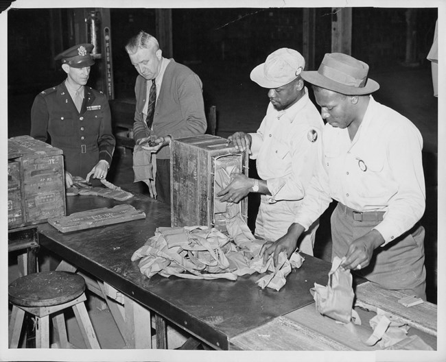 A white military officer inspects production as two Black male workers remove ammunition belts from boxes on a long table