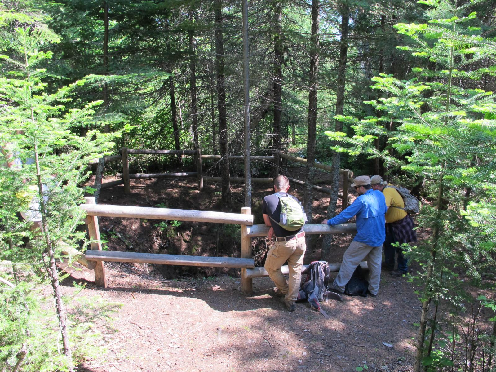 Three people lean on a wooden circular fence that surrounds a large hole in the ground. Conifer trees surround them.