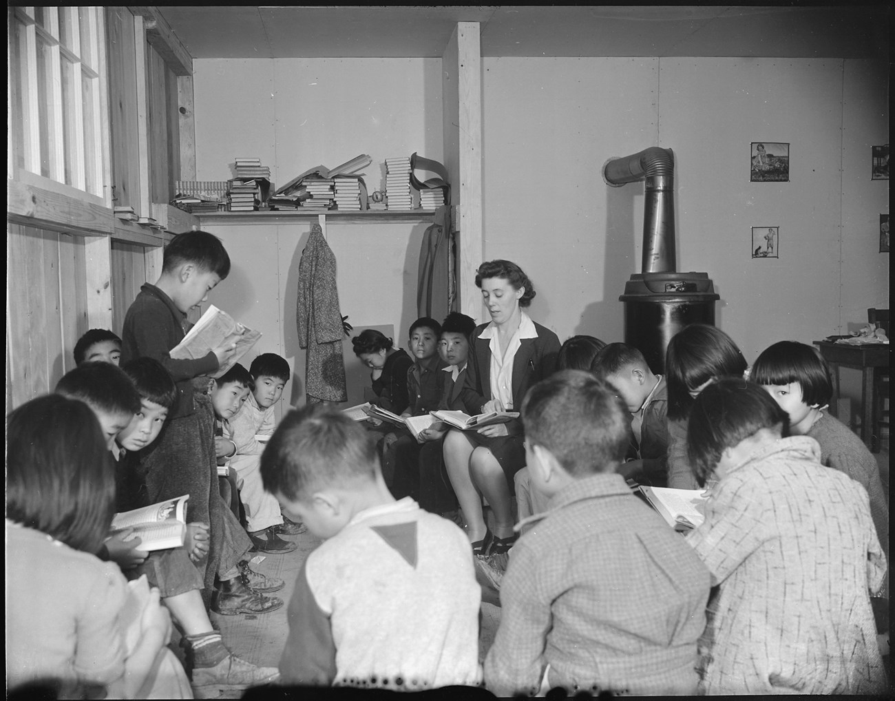Students of Japanese ancestry sit in a circle with books in their laps. On the left, one boy stands and seems to be reading from his book. Just right of center, a white woman sits in the circle and is reading her book. There is a pile of books, a few coat