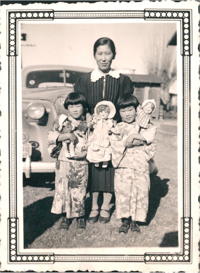 Black and white photo of a Japanese American woman and her two twin daughters. The woman wears a dark western-style dress. The girls wear Japanese kimonos and hold up their American baby dolls.